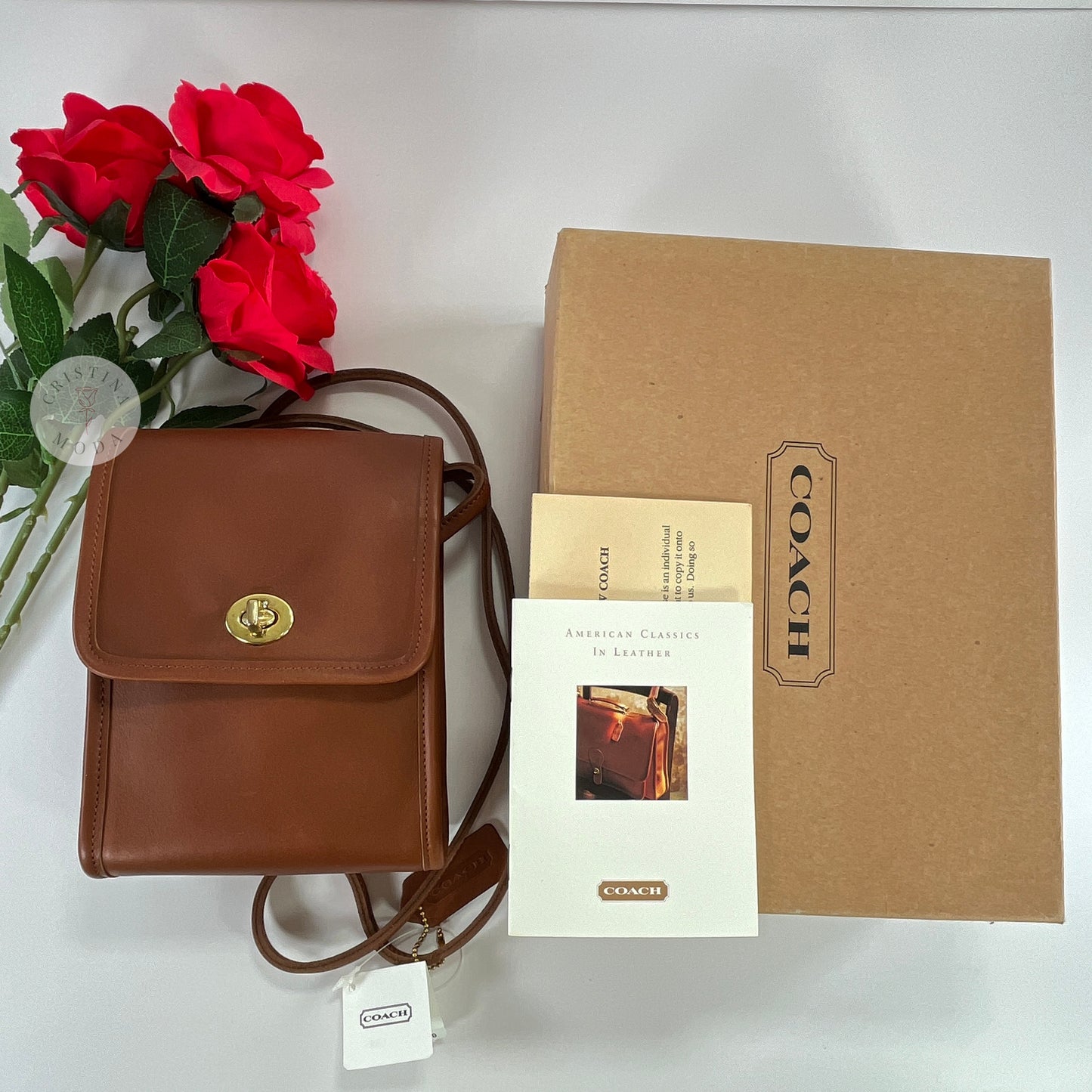 Top down view of the British Tan vintage Coach Scooter Bag laying flat on a white background with the box, booklet and registration card next to it and with 3 roses laying above it in the top left corner. 